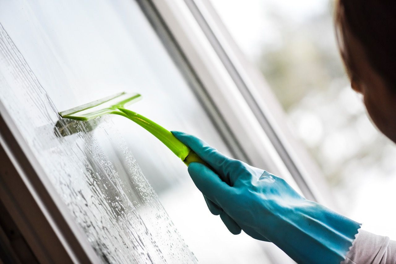 A hand wearing a blue rubber glove uses a green squeegee to clean a glass window pane.