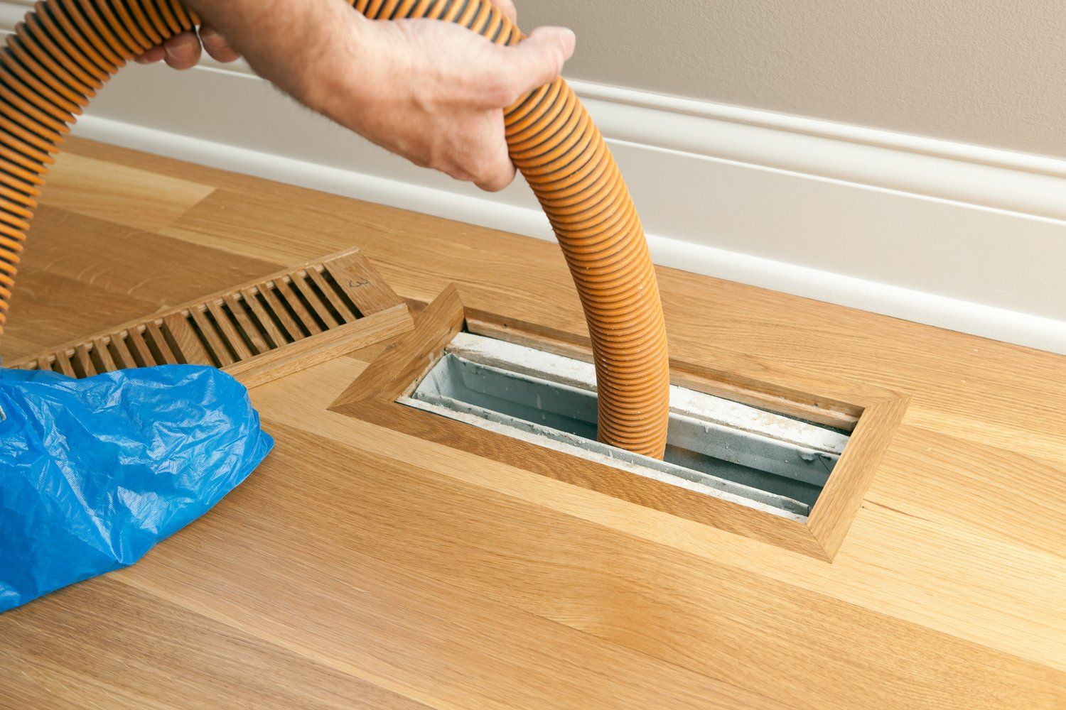 A person is using a vacuum hose to clean a vent on the floor.