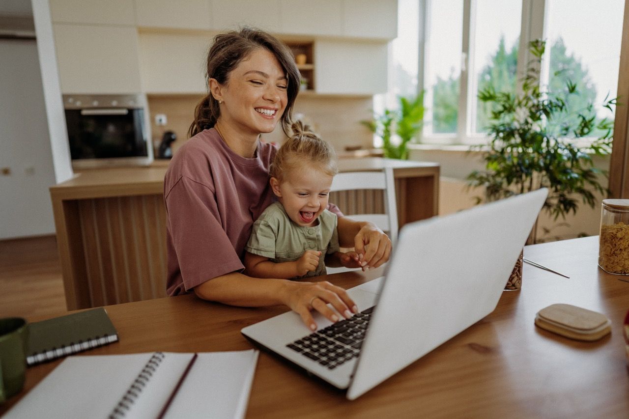 Woman smiles while using laptop with child on her lap in a kitchen. multi-function space in home.