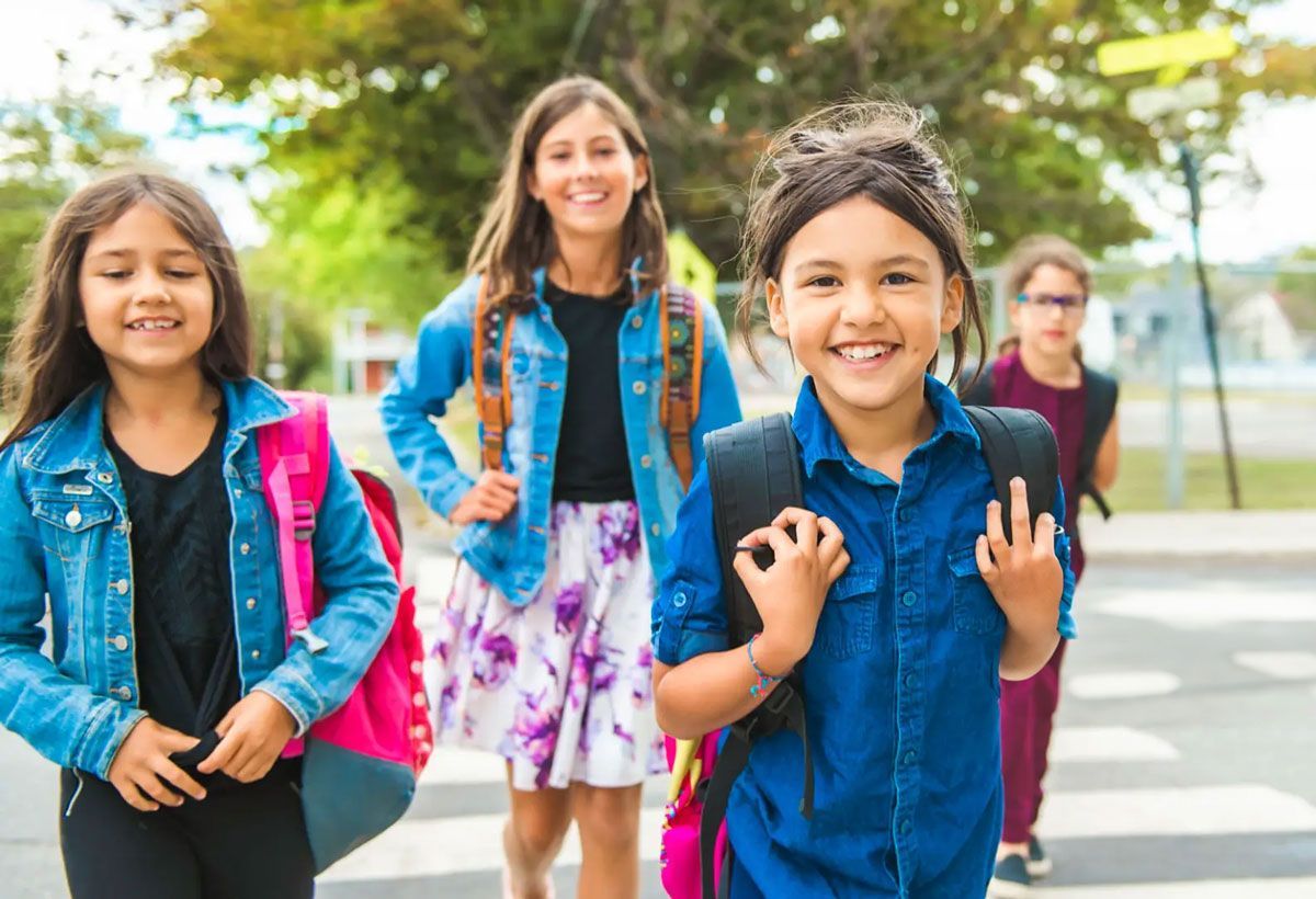 Group of smiling girls with backpacks walking to school, crossing a crosswalk outdoors. One girl makes an “okay” gesture.