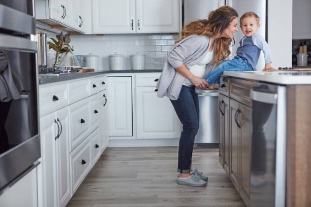 A woman is kneeling down in a kitchen with a child sitting on a counter.