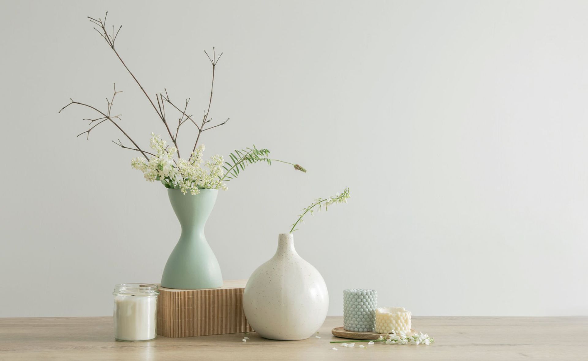 Two vases with floral branches, a small candle, and a textured white container sit on a wooden table against a white wall.