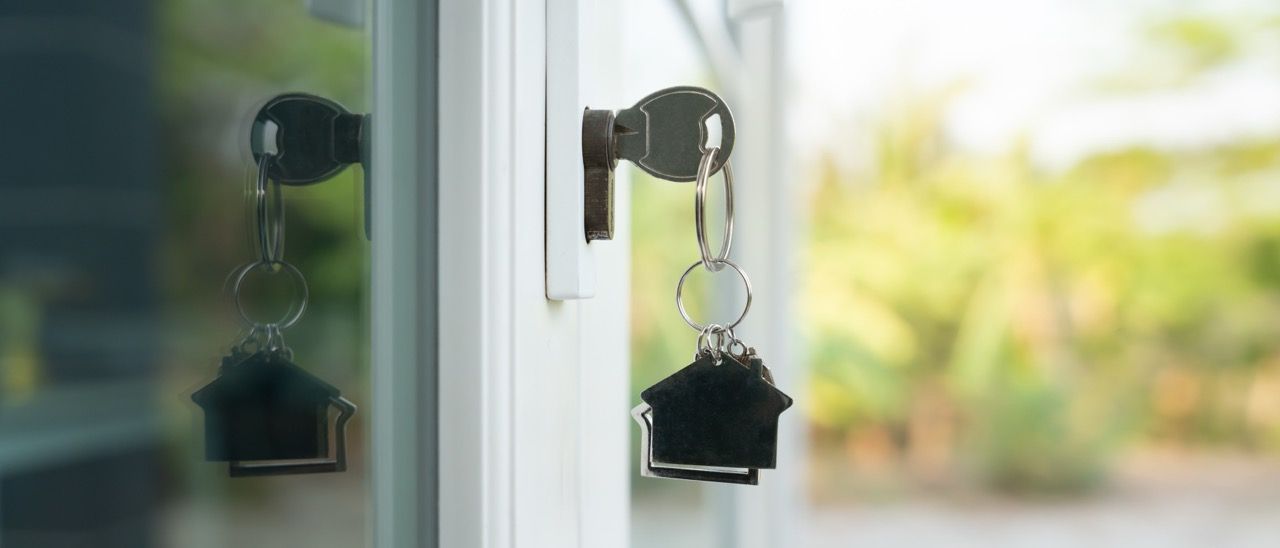 Keys in door, house-shaped keychain. Outside view of green trees, sunny day.