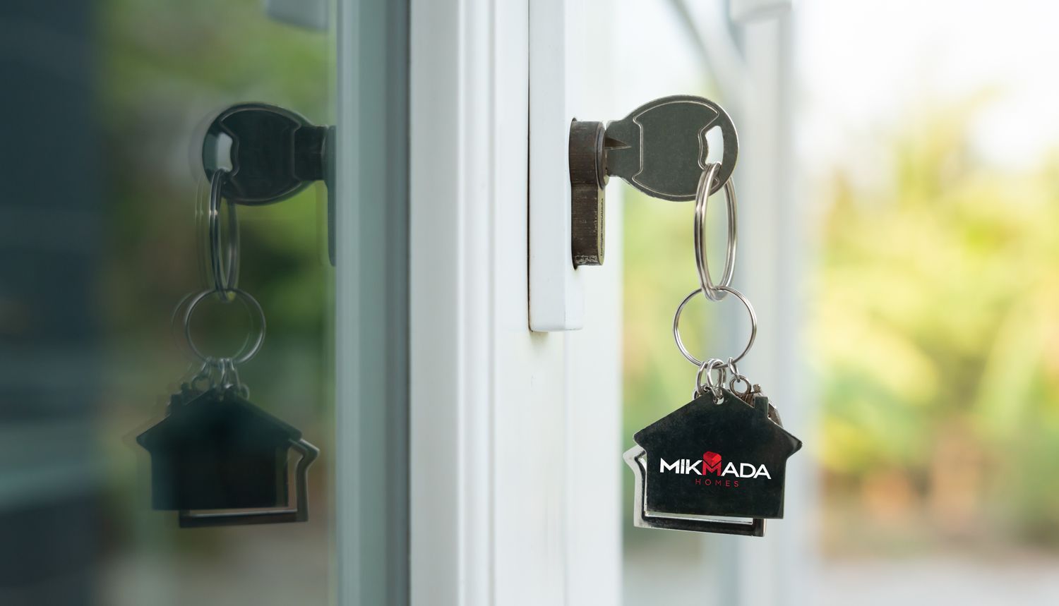 Keys in door, house-shaped keychain. Outside view of green trees, sunny day.