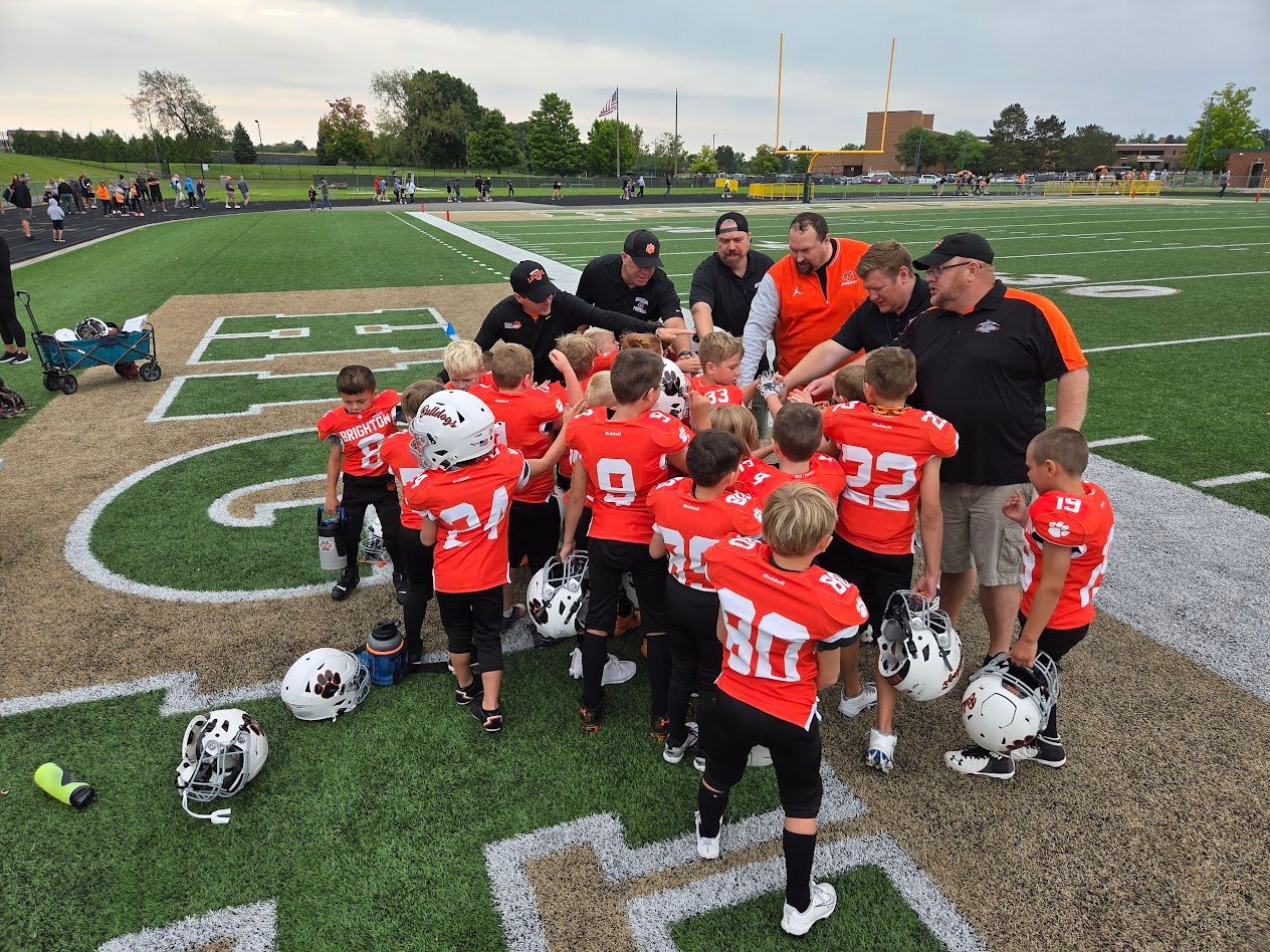 Youth football team in red jerseys huddles on a turf field with coaches after a game