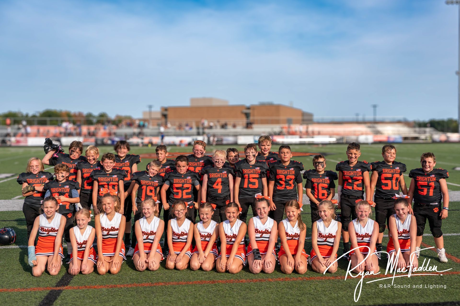 Youth football team posing on a field, players in black and orange uniforms with cheerleaders in front.