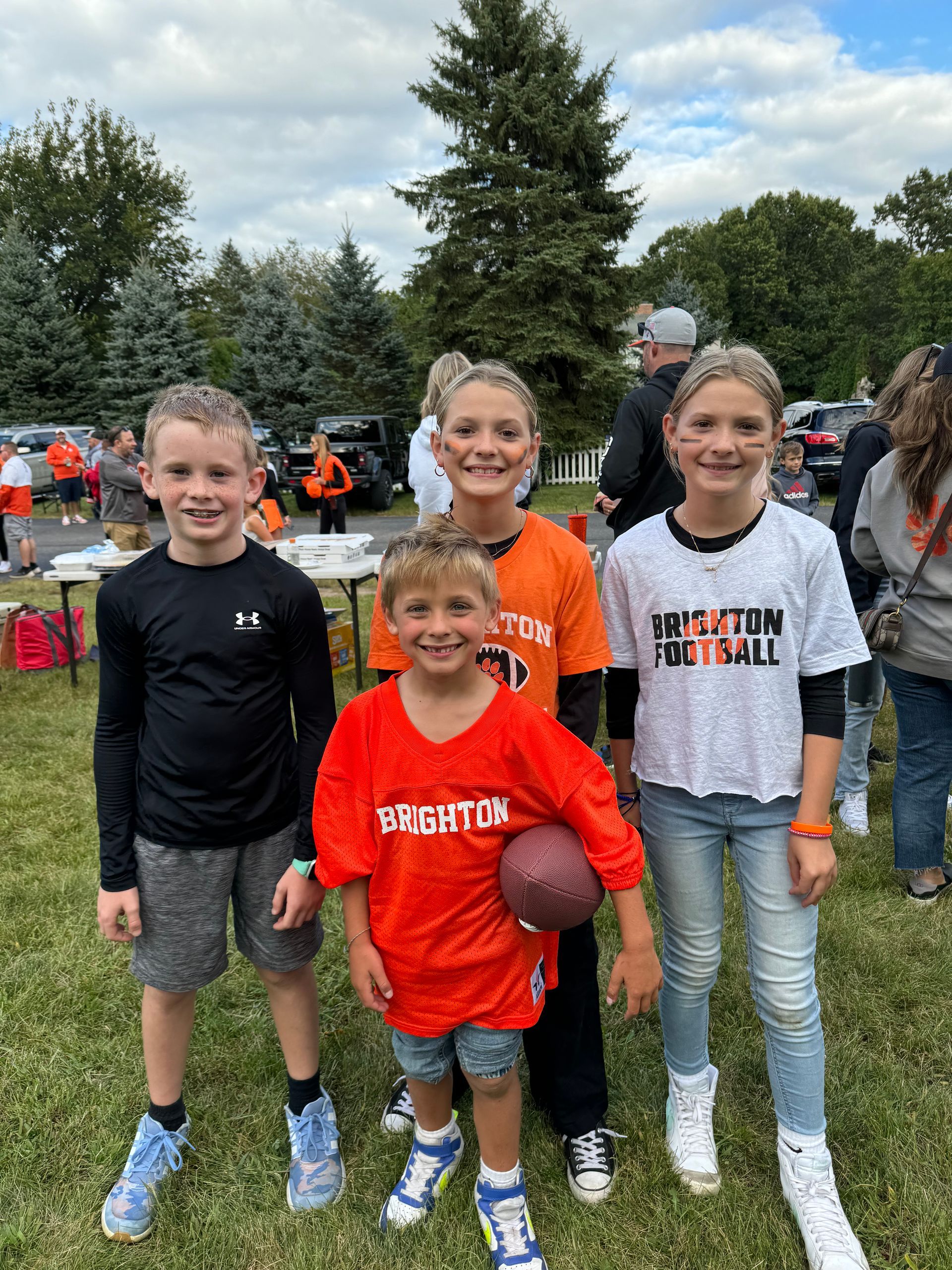 Four kids posing on a grassy field, two in orange shirts, with tents and people in the background.