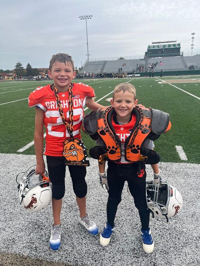 Two young football players in red and black uniforms stand on a field, smiling with helmets at their feet.