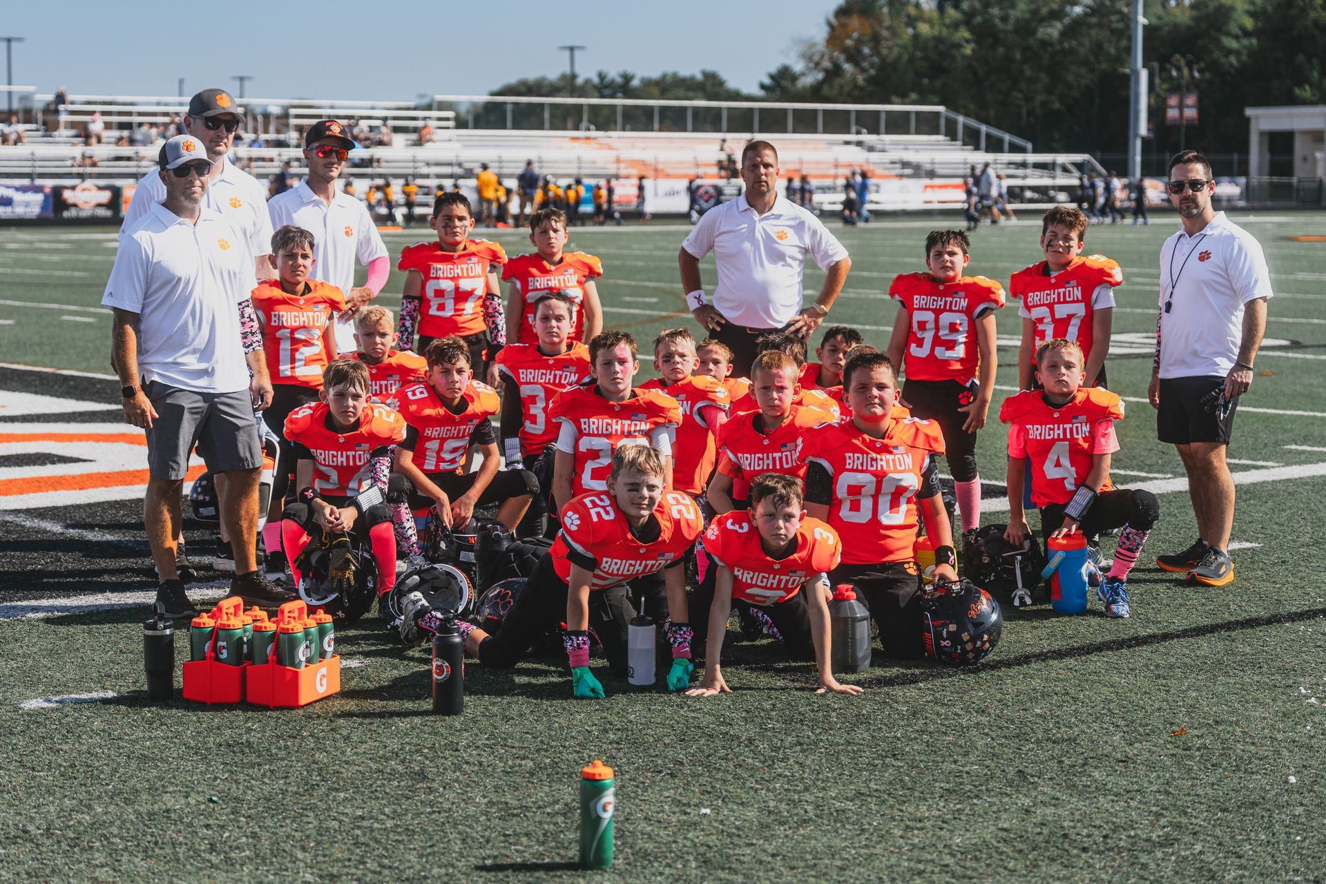 Youth football team and coaches posing on a field in red jerseys, with a trophy in front.