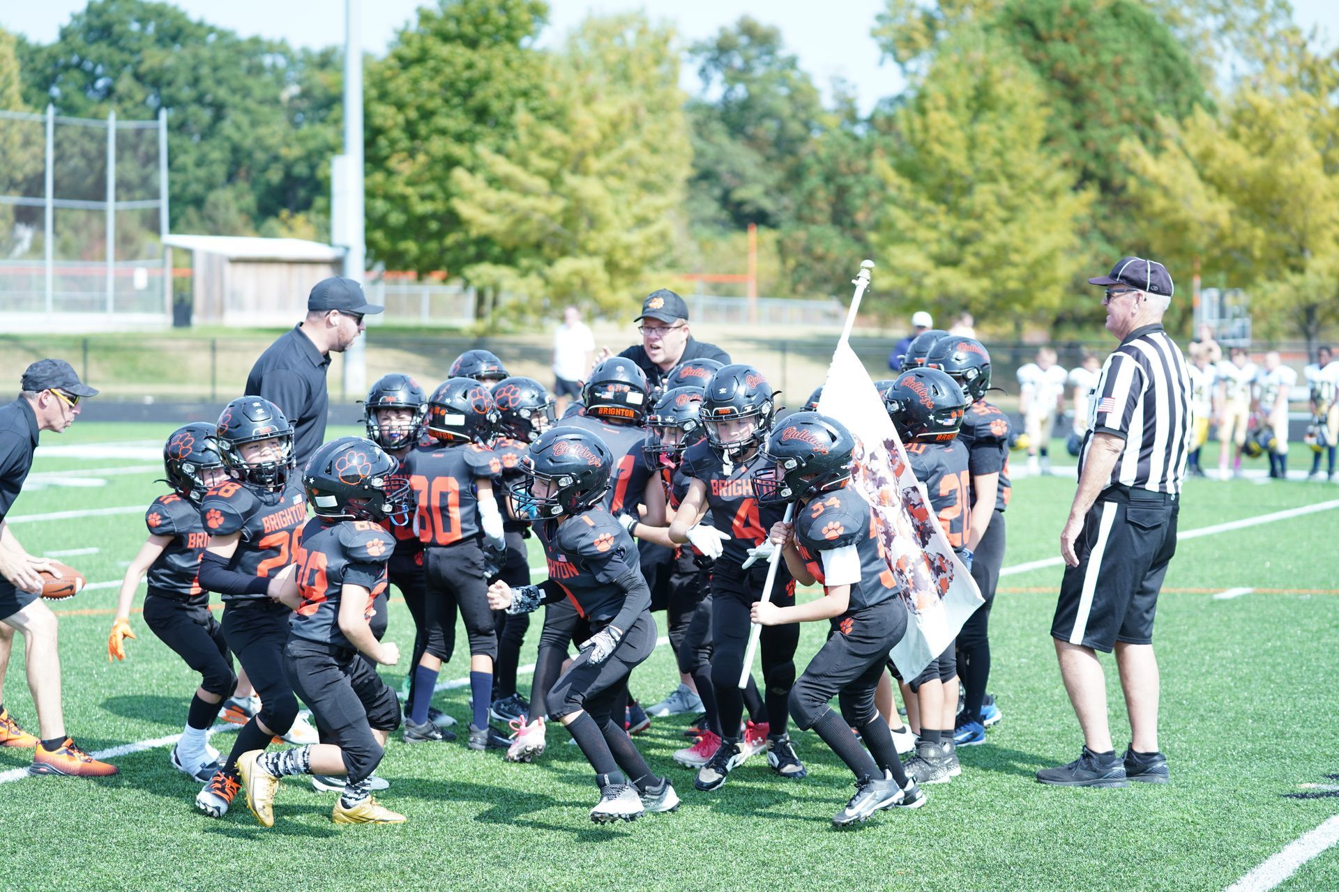 Youth football players in black uniforms scramble for the ball on a sunny field, with a referee nearby.