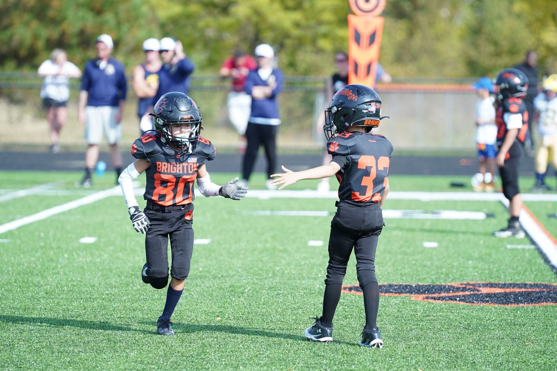 Two youth football players in black and orange uniforms on a field, one holding up a hand