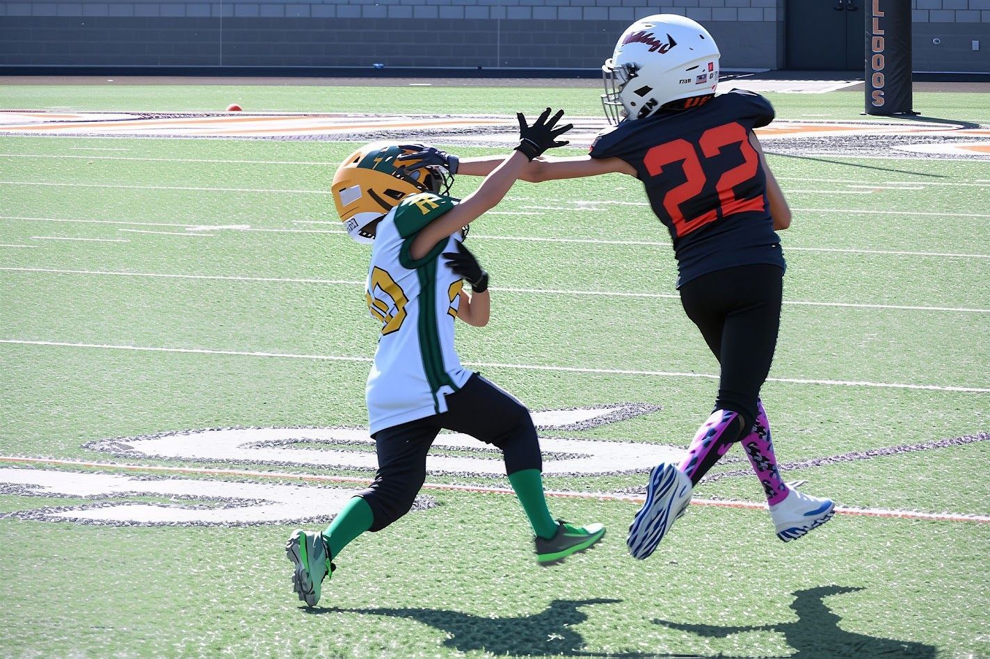 Two youth football players collide on a sunlit field, one in black and orange, the other in white, yellow, and green.