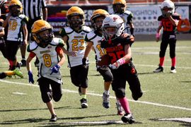 Youth football players running on a field, with a white-jersey player carrying the ball past black-and-orange defenders.