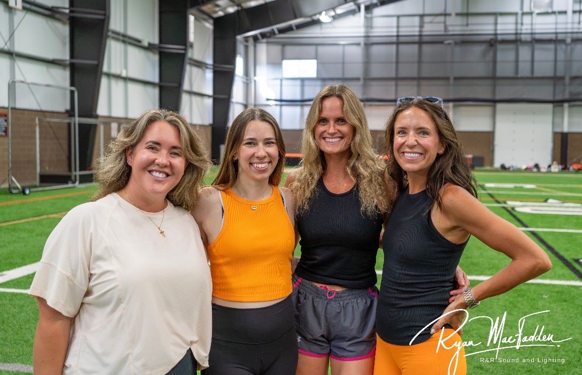 Four smiling people stand on an indoor turf field, posing in athletic wear.