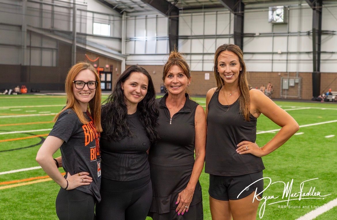 Four people pose smiling on an indoor soccer field, with a green turf and training facility in the background.