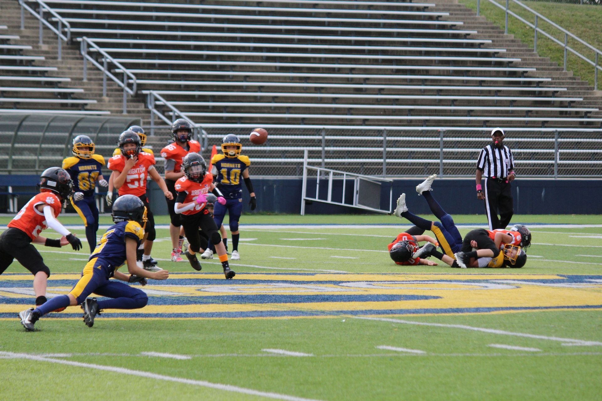 Youth football game on a field, with players tackling near the sideline and bleachers in the background