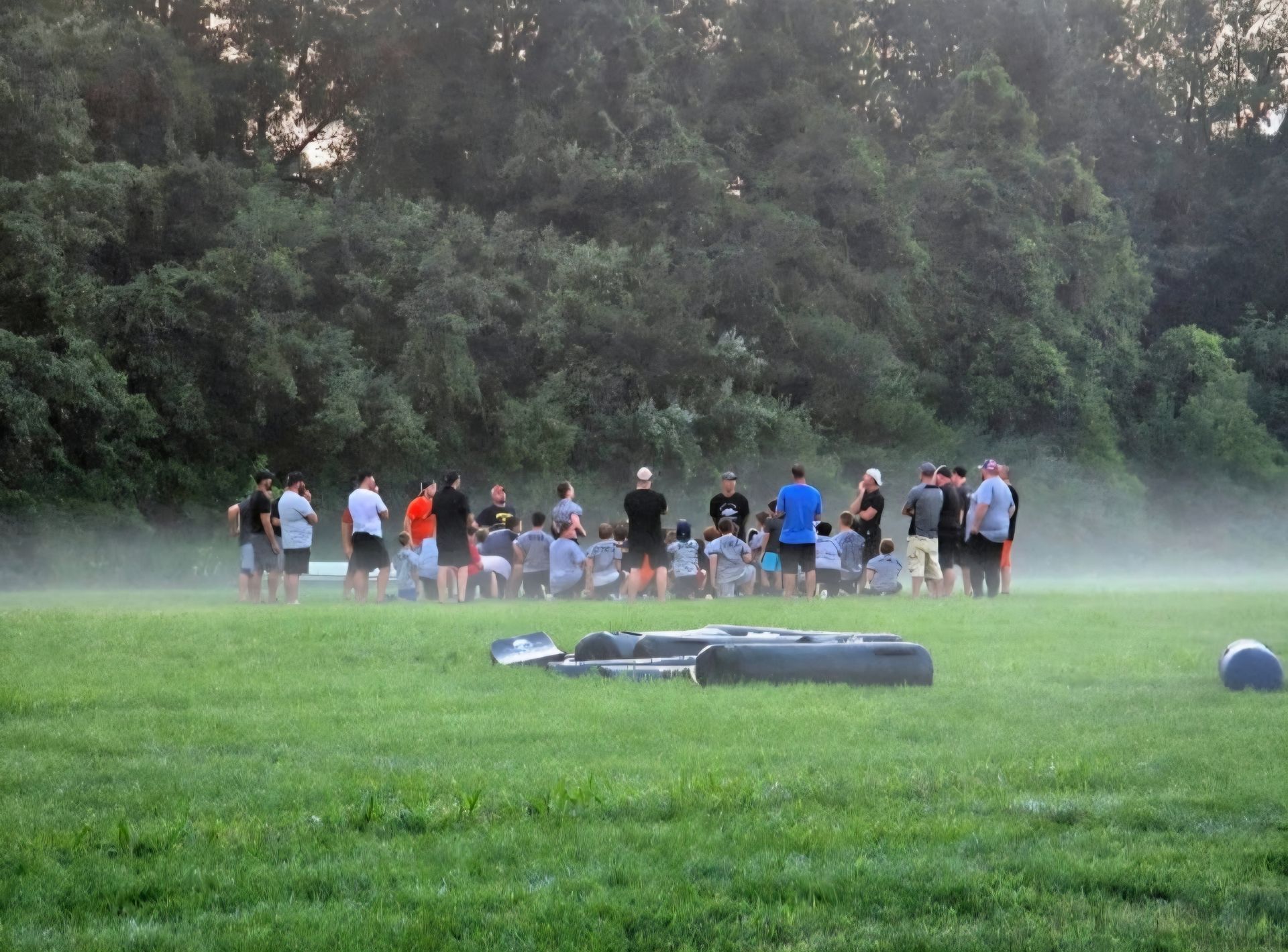 Group of people standing in a grassy field near a low obstacle, with mist and trees in the background
