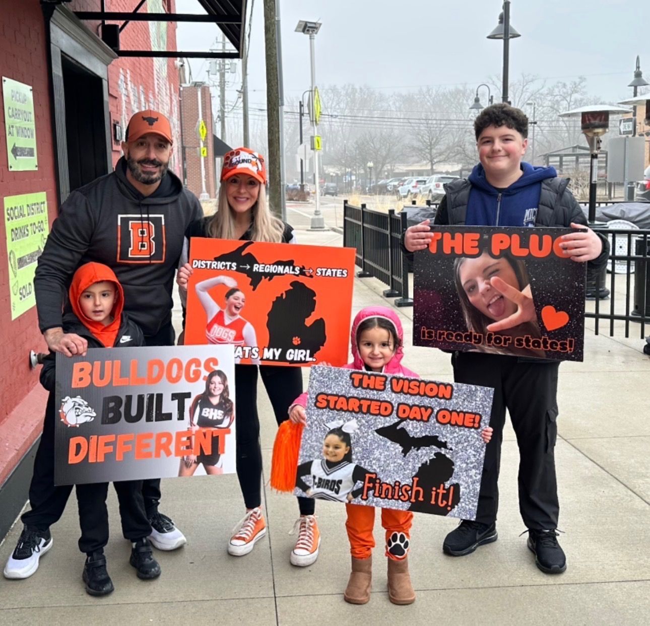 Family holding orange-and-black school spirit signs on a snowy sidewalk outside a storefront