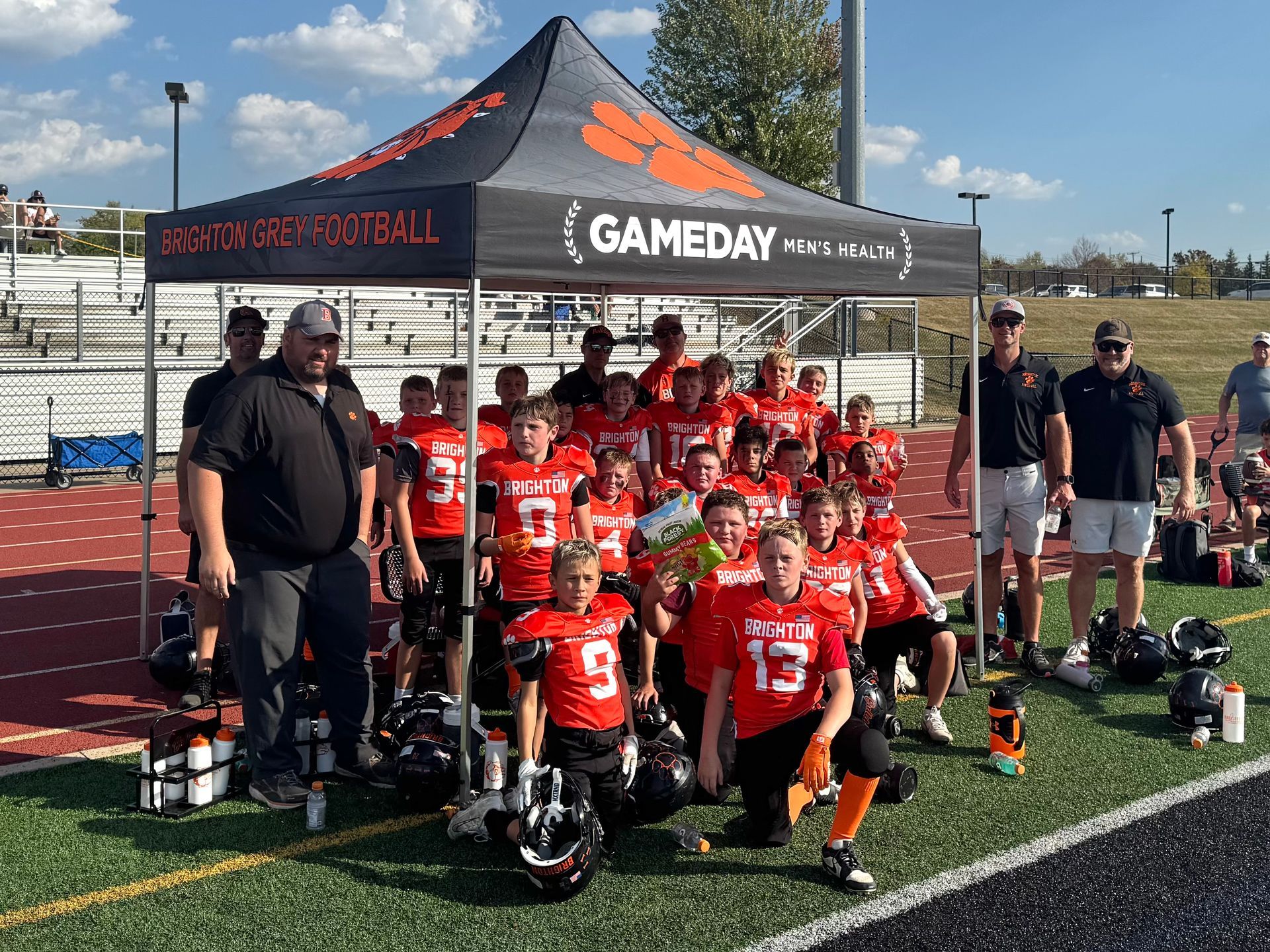 Youth football team posing under a black GAMEDAY canopy on a field, in red uniforms with coaches beside them
