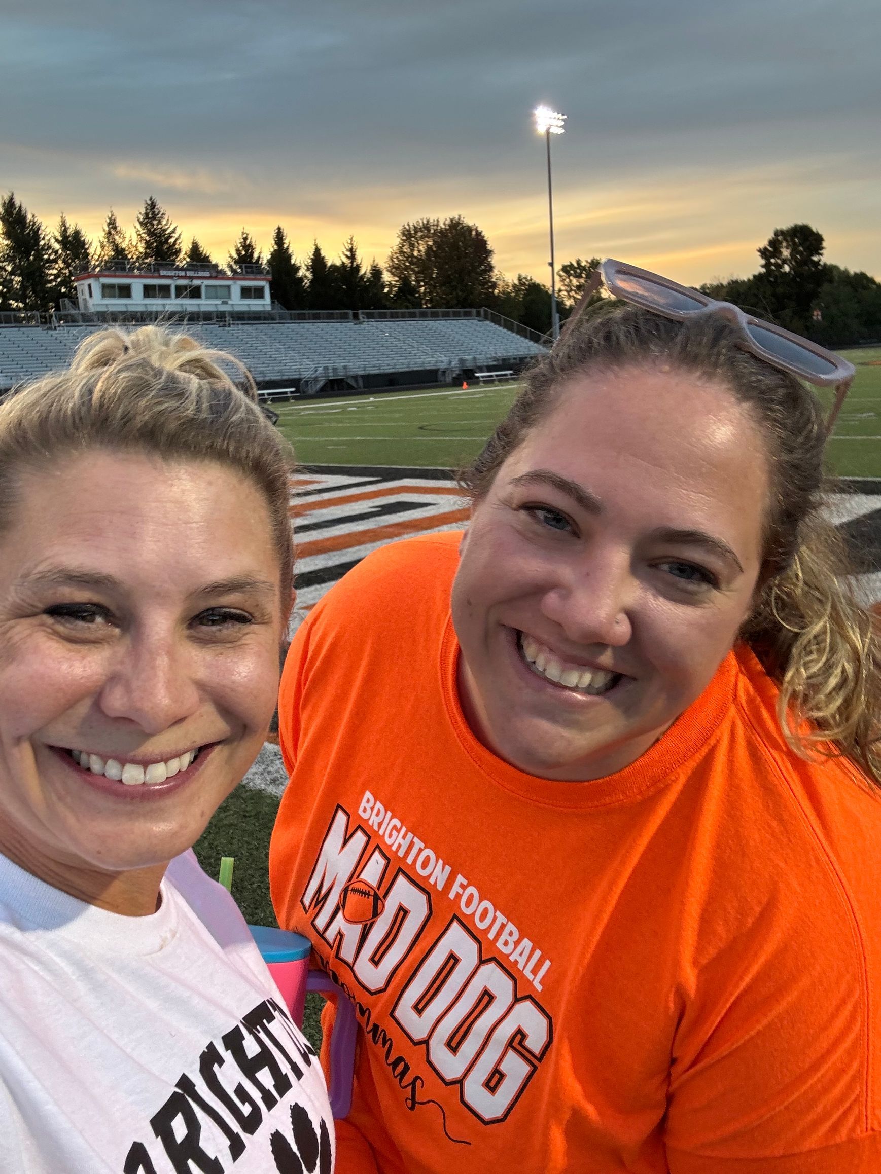Two smiling people taking a selfie at a football field at sunset, one in an orange shirt.