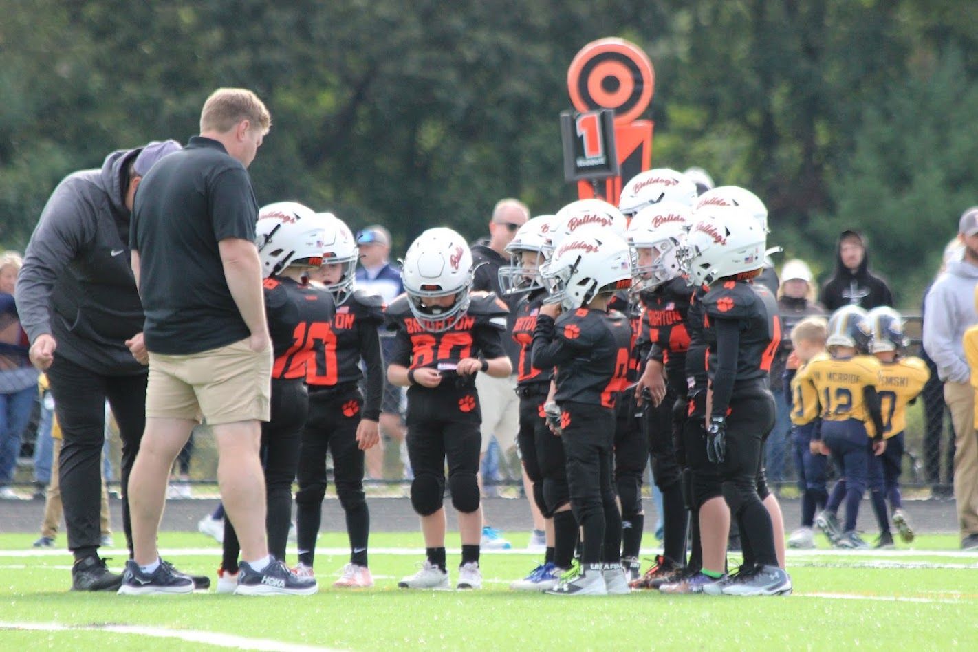Youth football team huddles with coach on the field, wearing black uniforms and white helmets under a down marker.