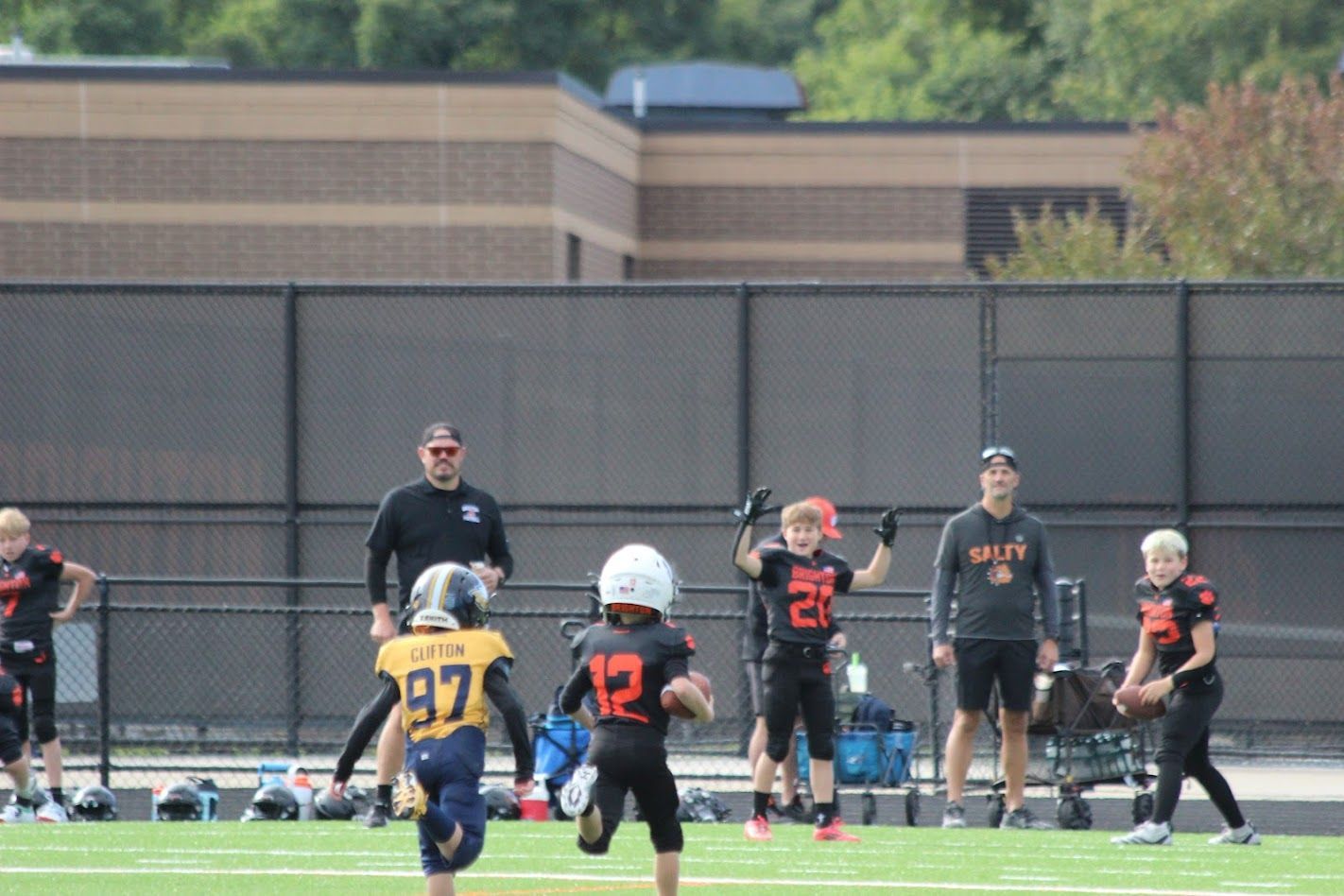 Youth football players and coaches on a field, with players in black and gold uniforms running play