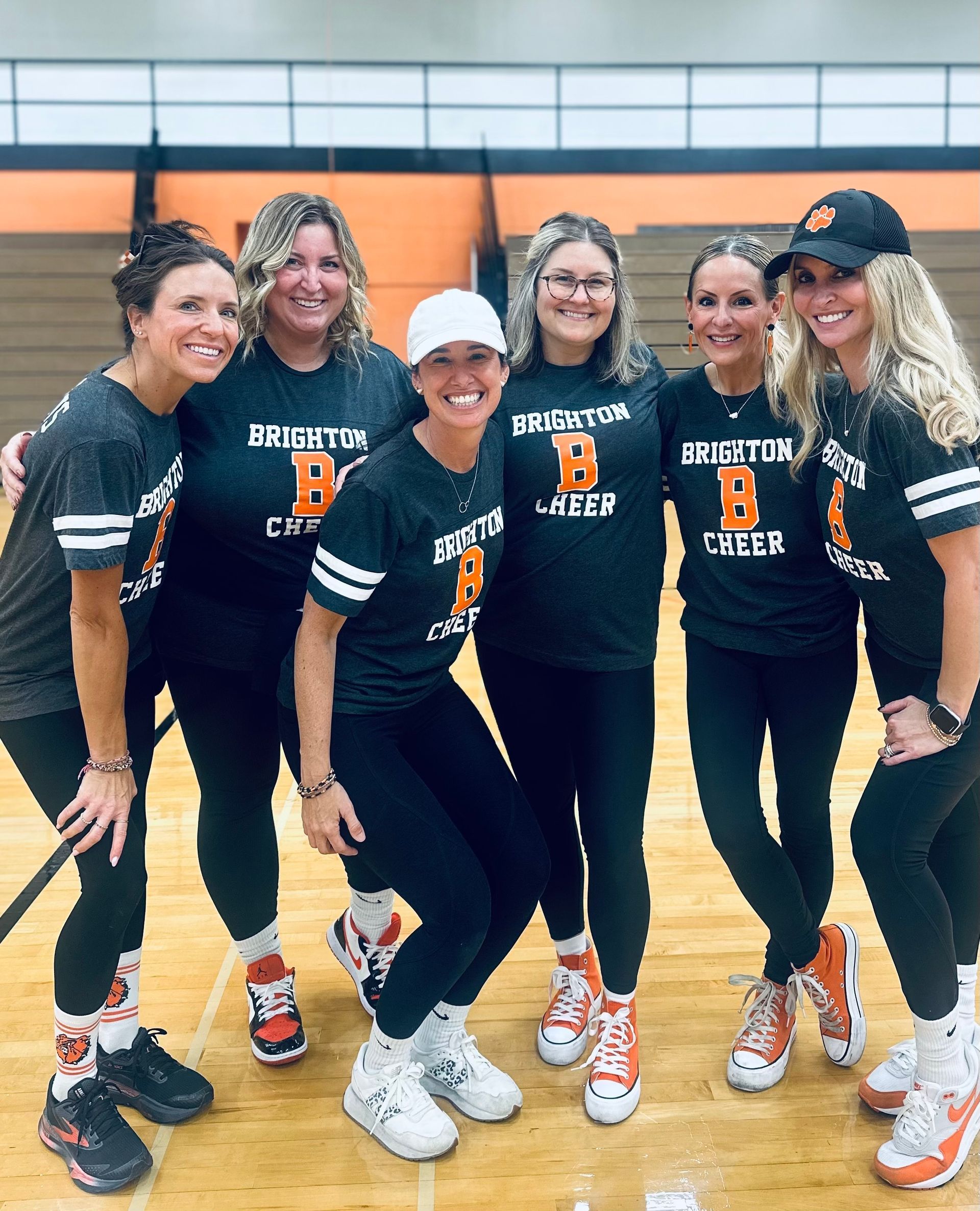 Six teammates in black volleyball uniforms pose smiling in a gym, one wearing a white cap.
