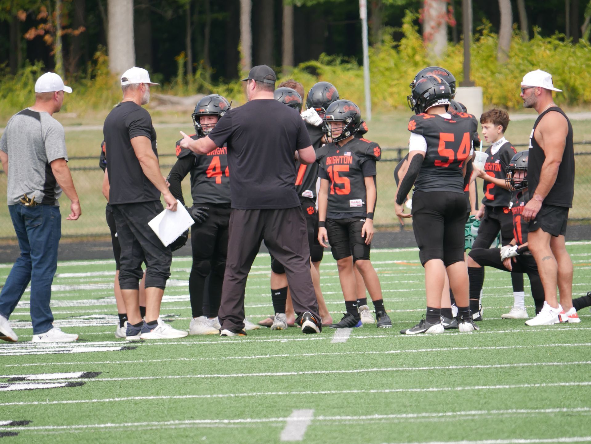 Football players and coaches huddle on a field during practice, some in black uniforms and white caps.