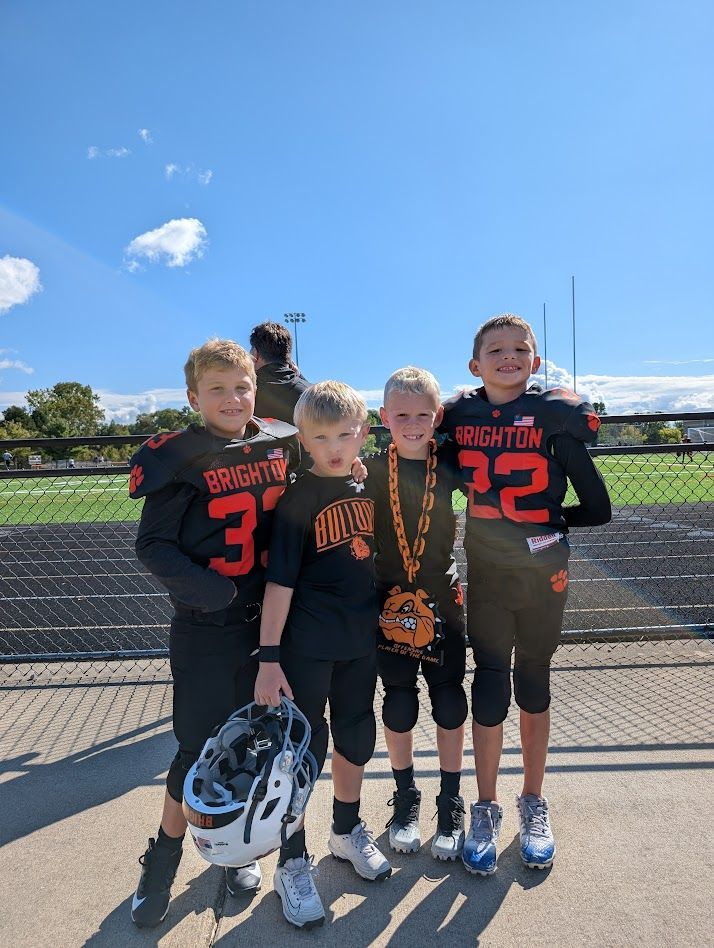 Four youth football players in black jerseys posing outdoors on a sunny field