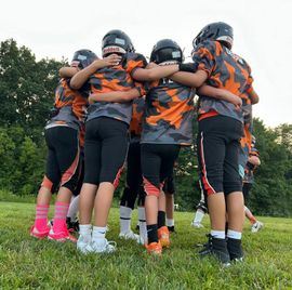 Youth sports team huddle on a grassy field, wearing black and orange uniforms and helmets.