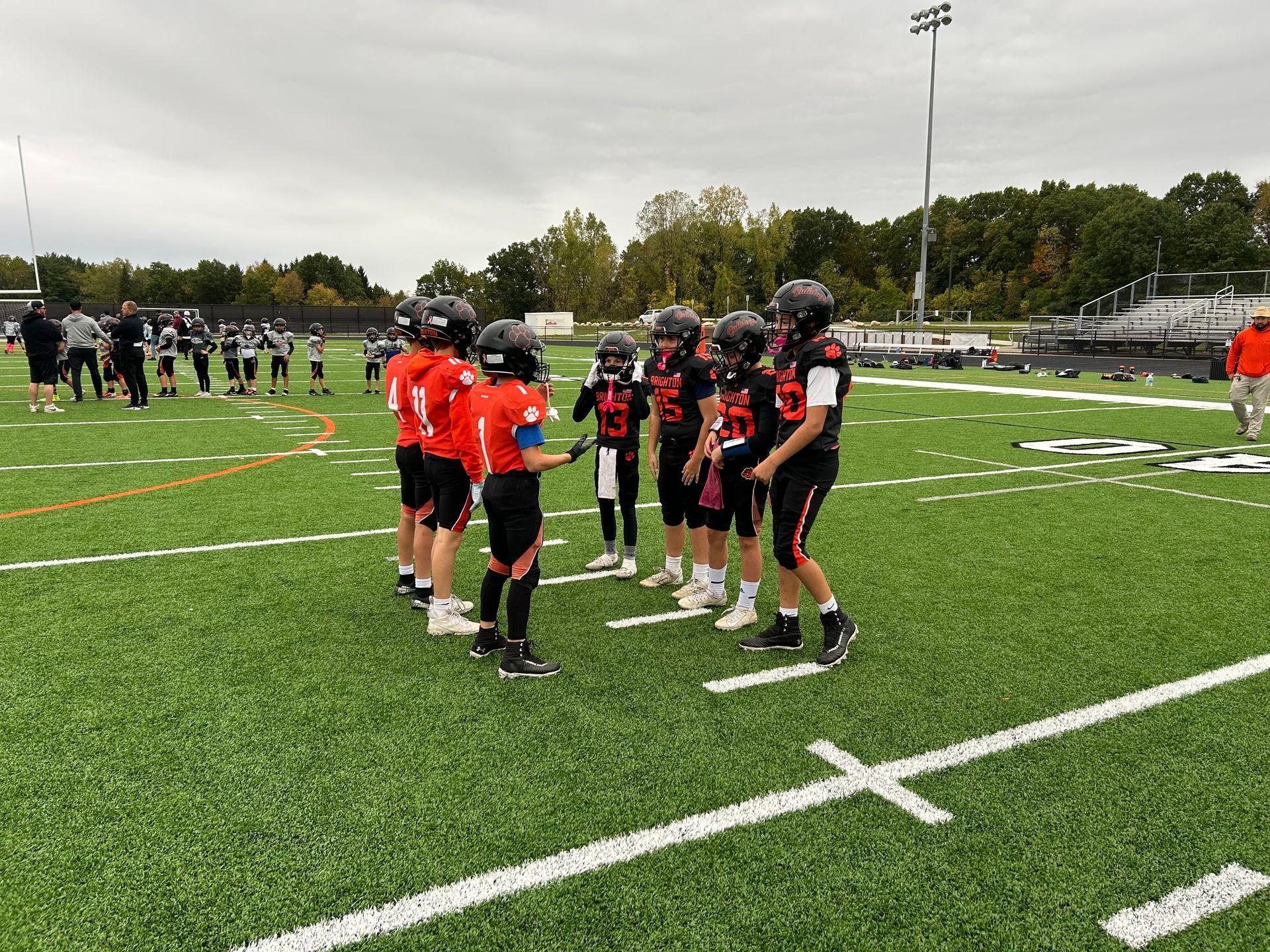 Youth football players in orange and black huddle on a field during practice.