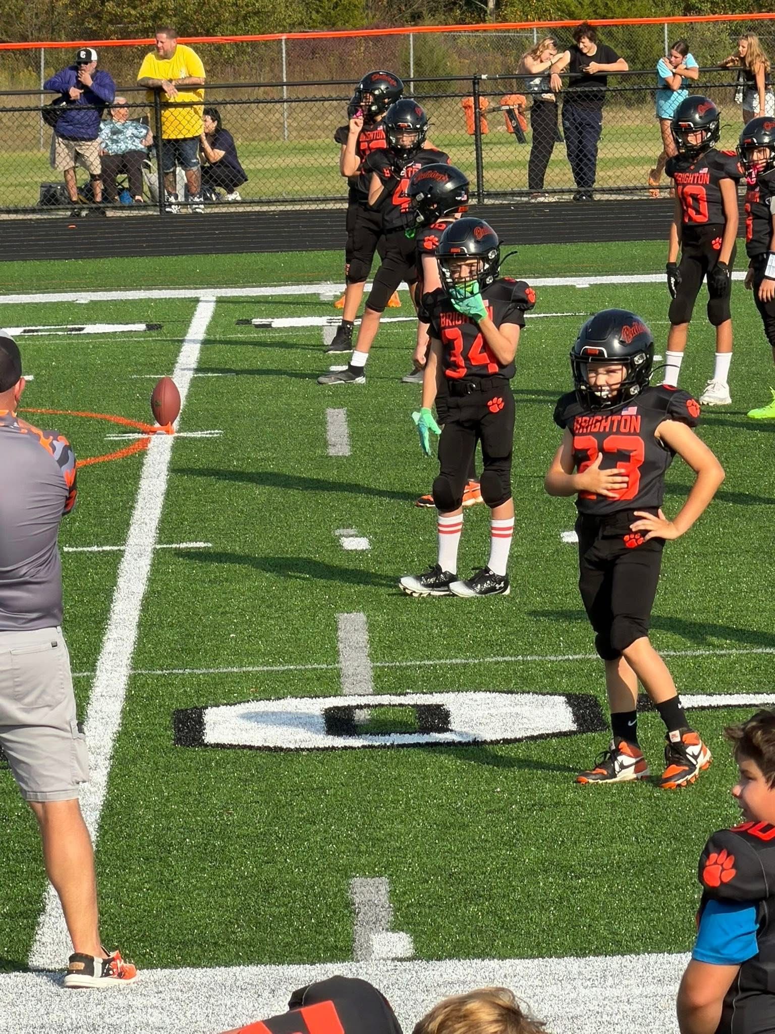Youth football players in black and orange uniforms lined up on a turf field near the sideline