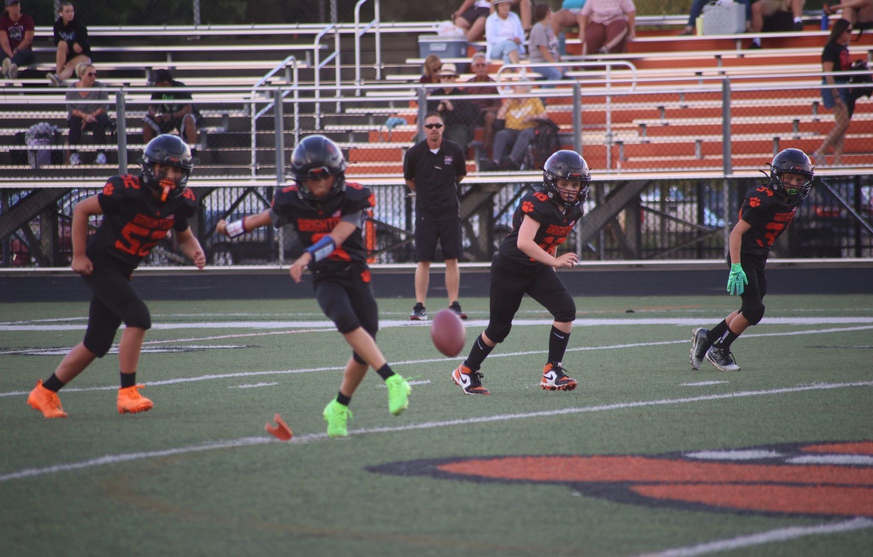 Youth football players sprint across a field during a game, with bleachers in the background.