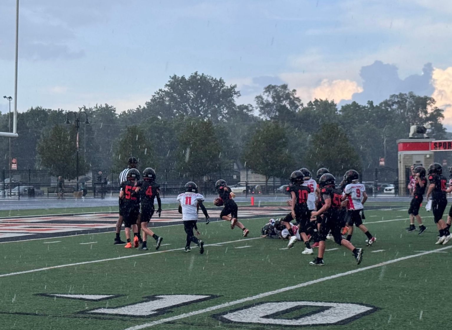 Youth football players in black uniforms on a field, gathered after a play near the 10-yard line at dusk.