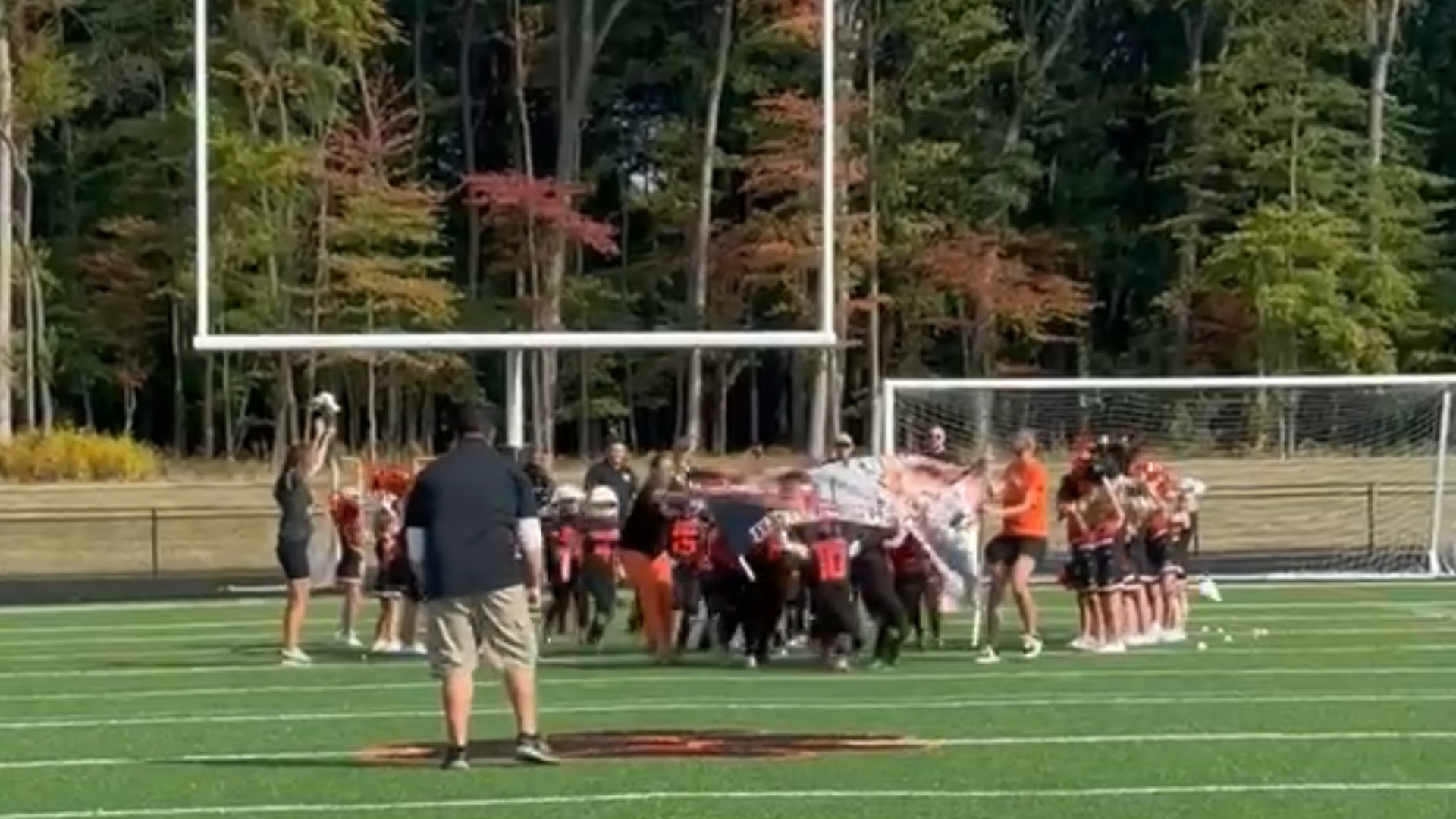 Football players huddle on a field near the goalpost, with coaches and trees in the background