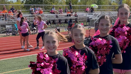 Cheerleaders in black uniforms with pink pom-poms stand on a track beside a football field.
