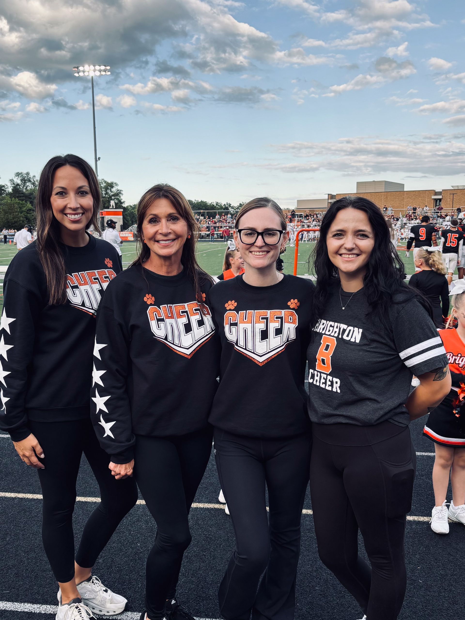 Four women in matching black event shirts pose on a football field with a crowd behind them.
