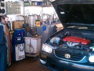 Car with hood open in auto repair shop; mechanic near equipment.