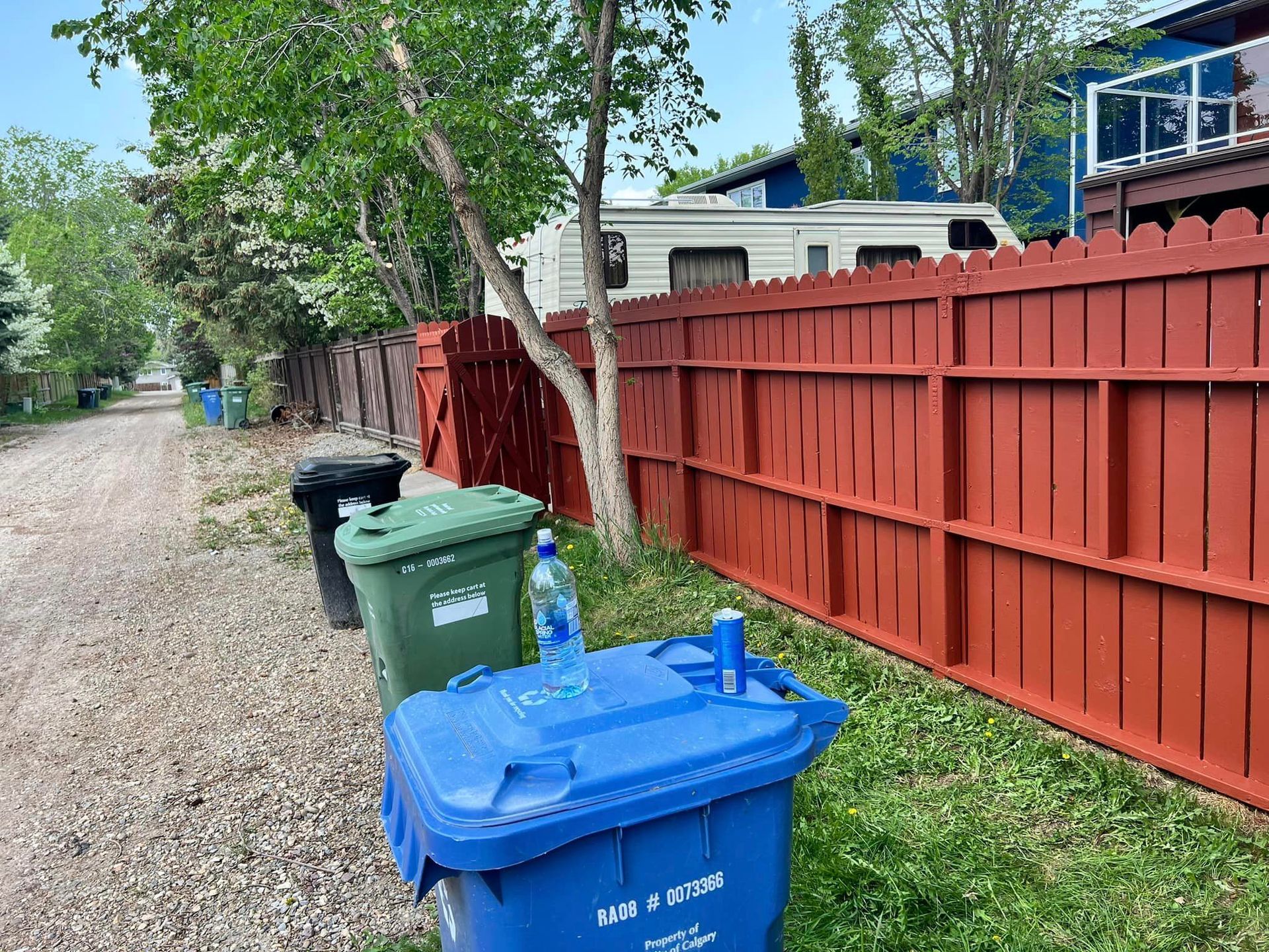 A blue trash can is sitting in front of a red fence.
