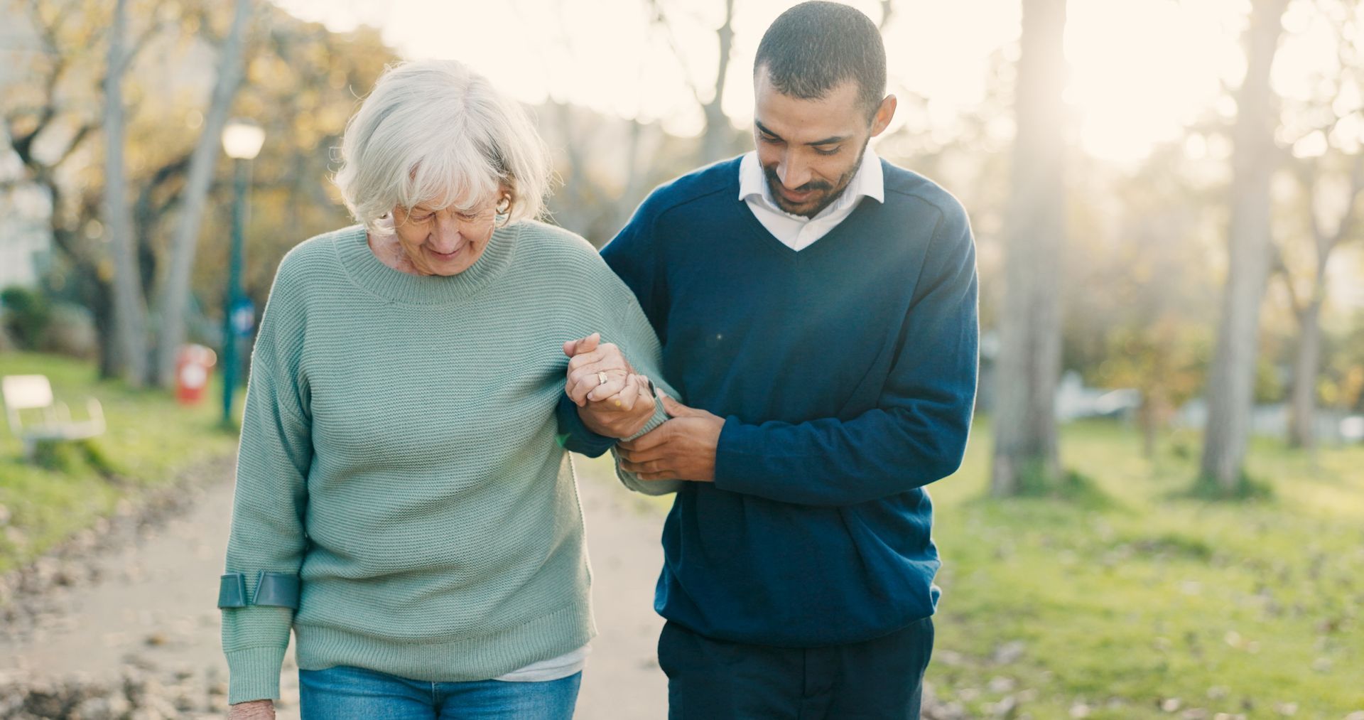 A man is helping an elderly woman walk in a park.