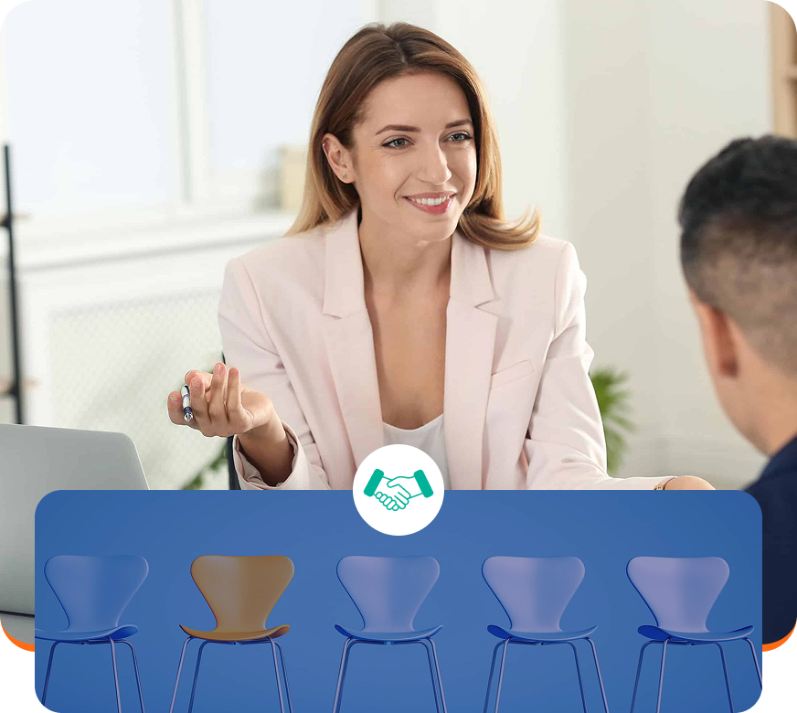 Woman in a pale pink blazer speaking at a table with blue chairs and a laptop in a bright office