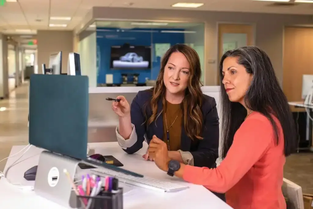 Two coworkers in an office discussing something at a desktop computer, one pointing at the screen.