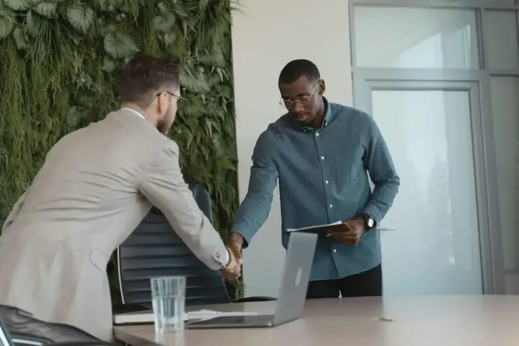 Two people shake hands across a table with a laptop in a modern office.