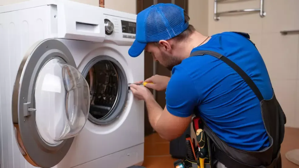 Man in blue overalls repairs a washing machine with a screwdriver.