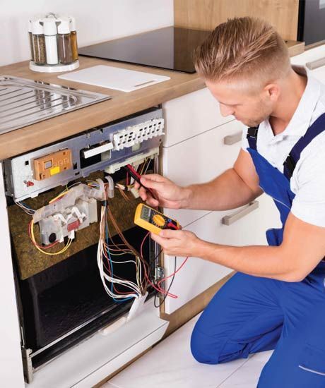 Man in blue overalls repairs a dishwasher with a multimeter in a kitchen.