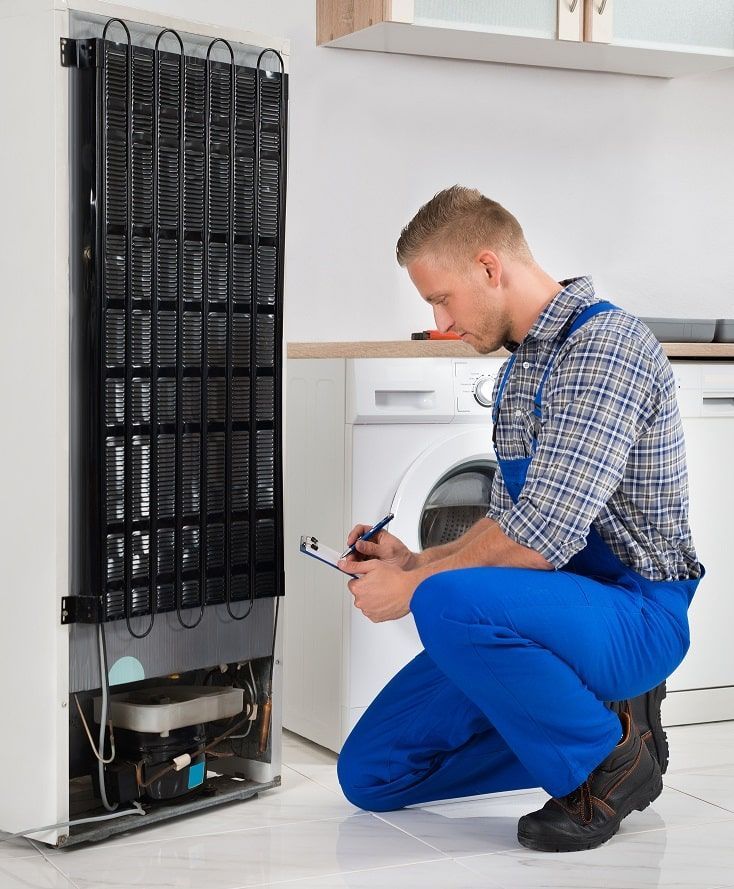 Man in blue overalls inspects a refrigerator, taking notes on a clipboard in a kitchen.