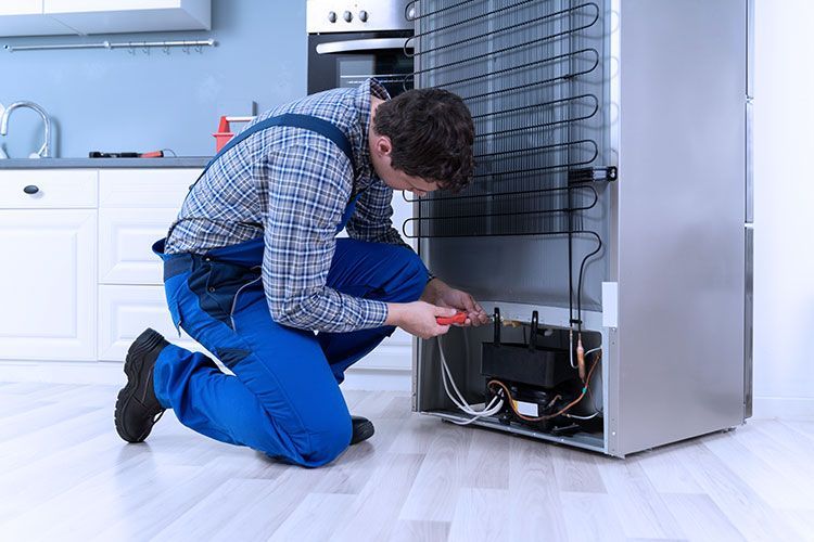 A repair person in blue overalls working on the back of a refrigerator in a kitchen.