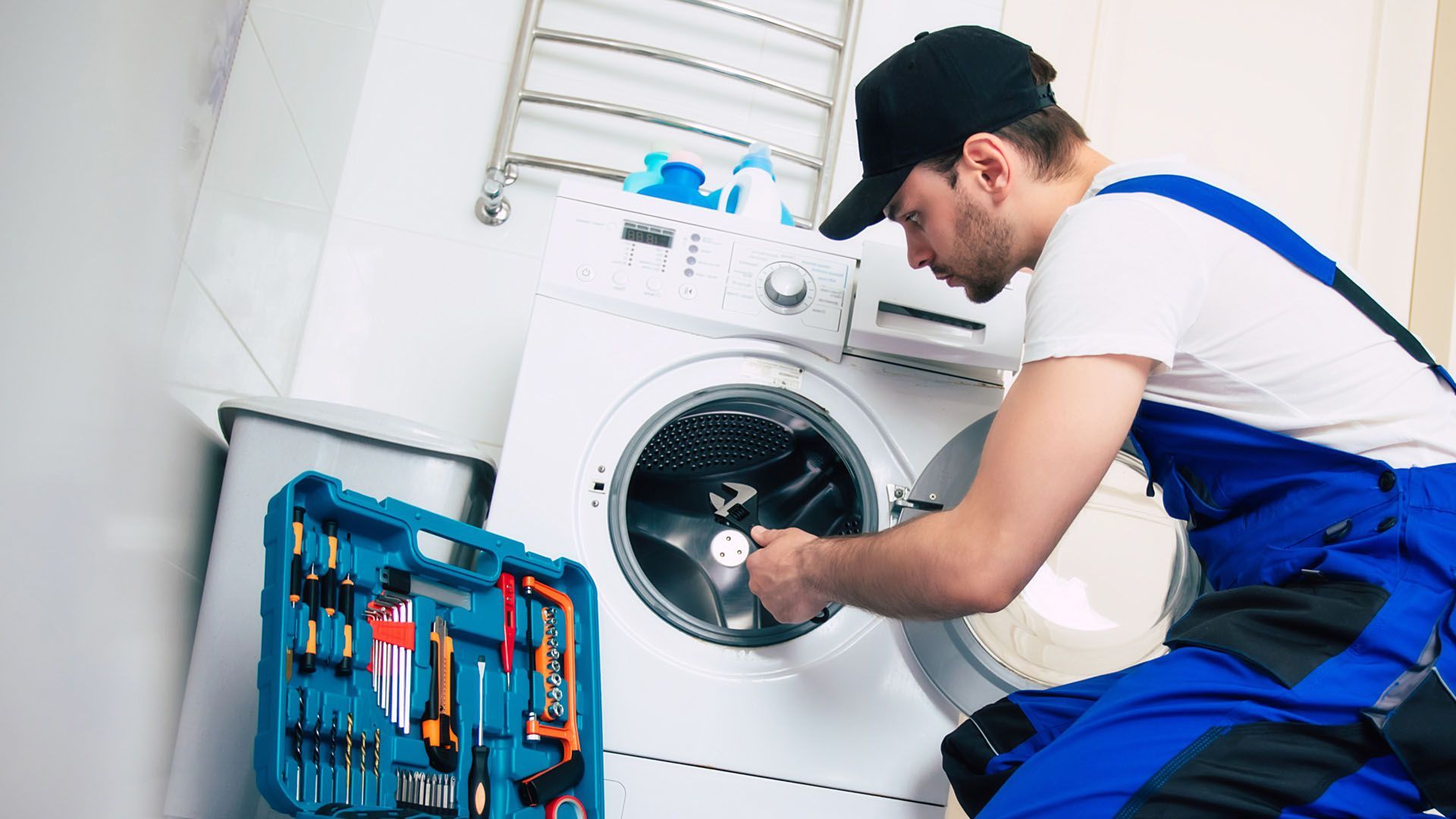 Man in blue overalls fixing a washing machine, toolbox nearby.