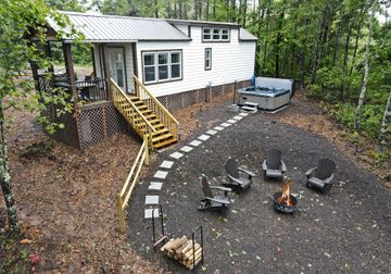 White cabin with yellow stairs overlooking a gravel fire pit area with chairs and wooded yard