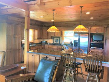 Warm log cabin kitchen and dining area with granite island, wooden chairs, pendant lights, and a view of the deck.